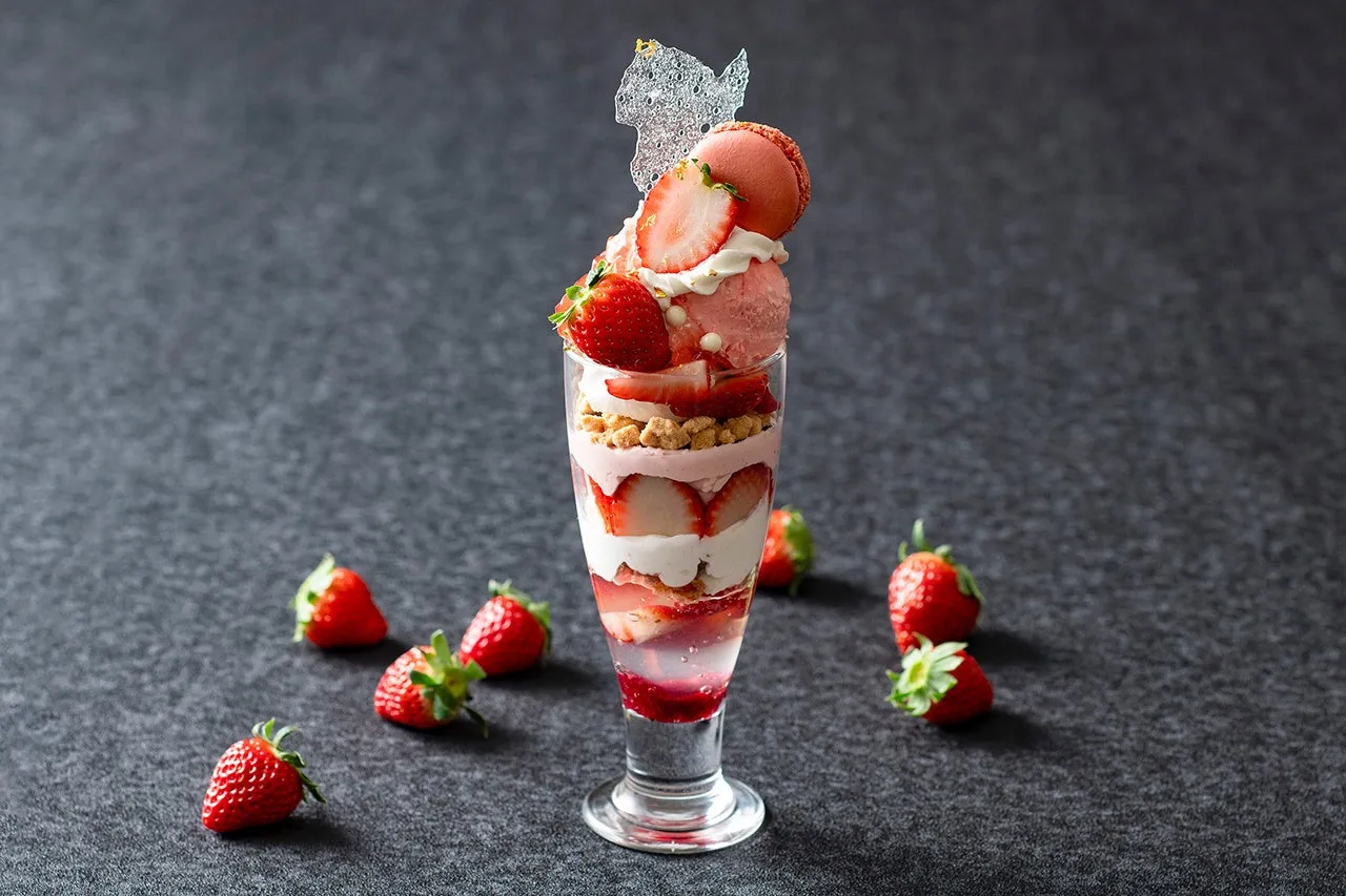 グラスに入ったイチゴパフェの写真。パフェは、イチゴ、クリーム、クランブル,アイスクリーム,マカロンなどで構成されている。周りにはイチゴが散りばめられている。