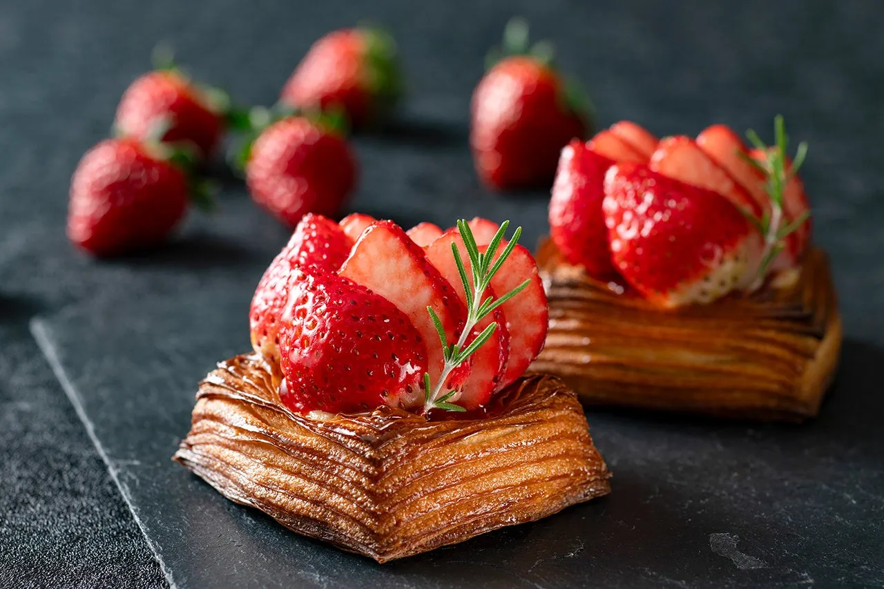 いちごがトッピングされたデザートのクローズアップ写真。デザートは四角いパイ生地の上にいちごが乗っており、背景にはぼやけたイチゴがいくつか写っています。