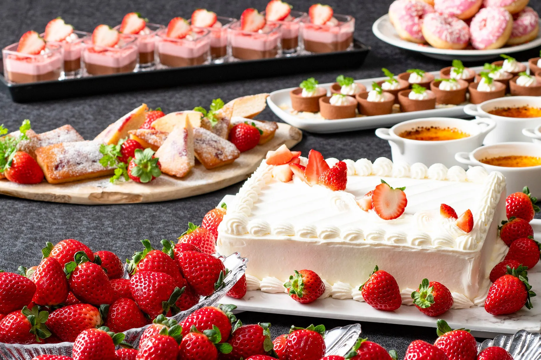 様々なデザートが並んだ写真。イチゴをふんだんに使ったケーキ、プリン、ドーナツ、タルトなど、様々な種類のデザートがテーブルいっぱいに並べられている。